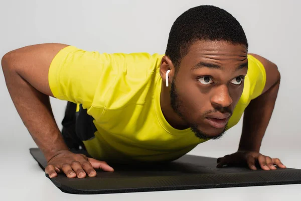 Sportive african american man in earphones doing push ups on fitness mat isolated on grey — Stock Photo