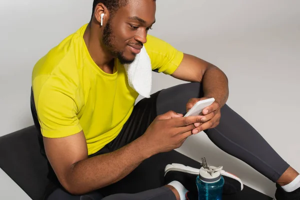 Zufriedener afrikanisch-amerikanischer Sportler sitzt auf Fitnessmatte und benutzt Smartphone in der Nähe von Sportflasche isoliert auf grau — Stockfoto