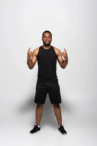 Comprimento total de Africano americano desportista em top tanque e shorts mostrando sinal de rocha em cinza — Fotografia de Stock