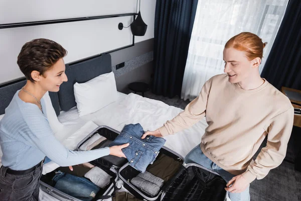 Vue grand angle du couple souriant tenant des vêtements près des valises sur le lit dans la chambre d'hôtel — Photo de stock