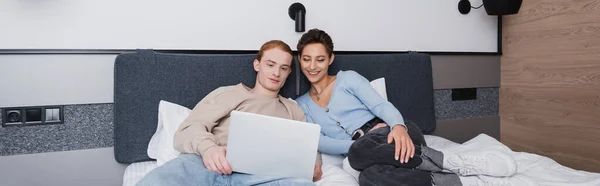 Donna sorridente sdraiata sul letto vicino al fidanzato con laptop in camera d'albergo, banner — Foto stock
