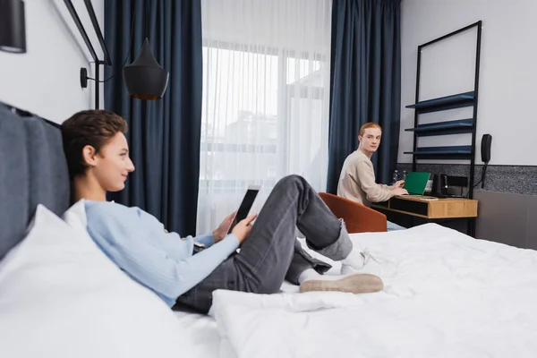 Jovem usando laptop perto de namorada borrada com tablet digital no quarto de hotel — Fotografia de Stock