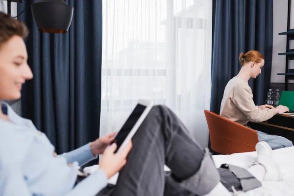 Side view of man using laptop near blurred girlfriend with digital tablet in hotel room — Stock Photo