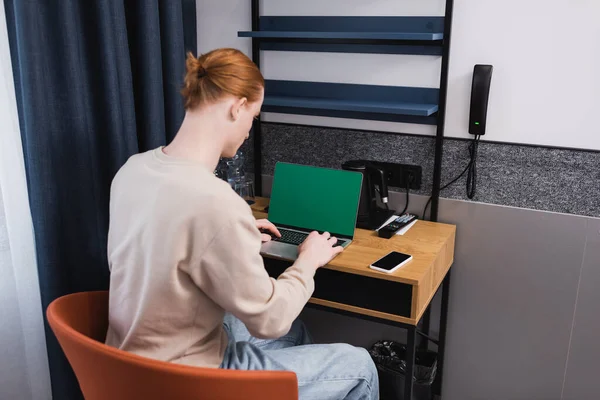 Redhead hombre usando el ordenador portátil con pantalla verde cerca de teléfono inteligente en la habitación del hotel - foto de stock