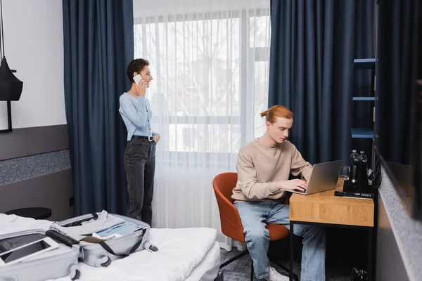 Улыбающаяся женщина разговаривает на смартфоне рядом с парнем с помощью ноутбука и чемодана на кровати в отеле — стоковое фото