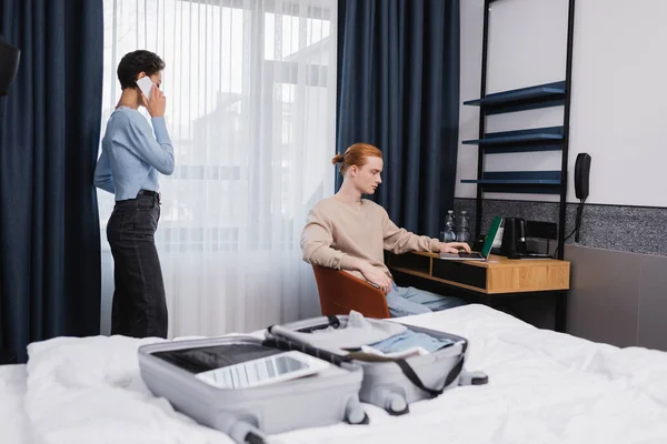 Hombre joven usando el ordenador portátil cerca de novia hablando en el teléfono inteligente y la maleta borrosa en la habitación de hotel - foto de stock