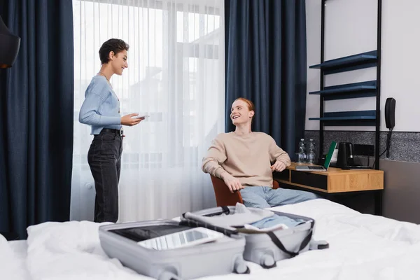 Positive young couple with gadgets talking near suitcase on bed in hotel room — Fotografia de Stock