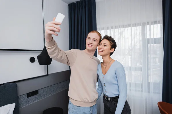 Couple souriant prenant selfie sur smartphone dans une chambre d'hôtel moderne — Photo de stock