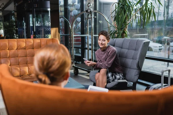 Smiling woman with cellphone looking at blurred boyfriend in hotel lobby — Stock Photo