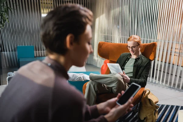 Young man using digital tablet near blurred girlfriend with smartphone in hotel lobby — Stock Photo