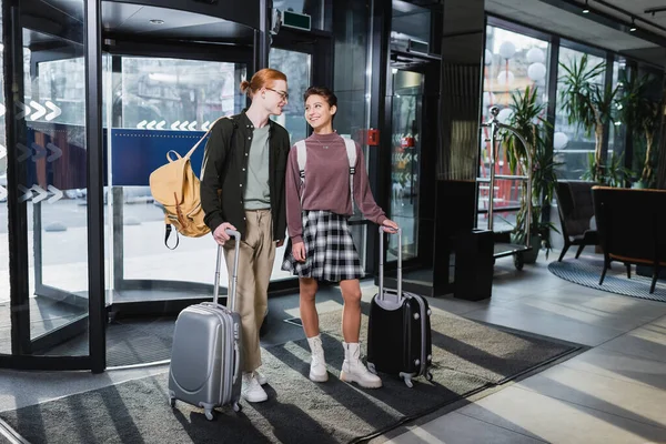 Smiling woman with backpack and suitcase standing near boyfriend in hotel lobby — стоковое фото