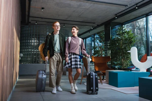 Cheerful woman with backpack and suitcase walking near boyfriend in hotel lobby — стоковое фото