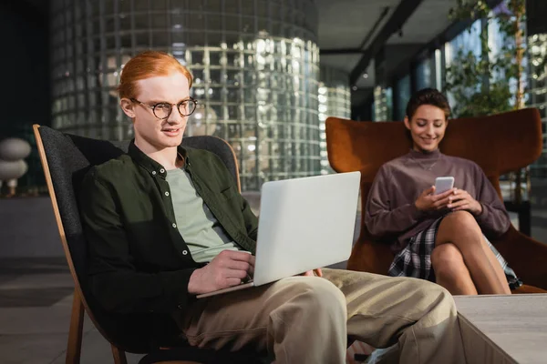 Smiling freelancer using laptop near blurred girlfriend with smartphone in hotel lobby — Stock Photo