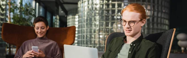 Young man looking at laptop near blurred girlfriend with smartphone in hotel lobby, banner — Stock Photo