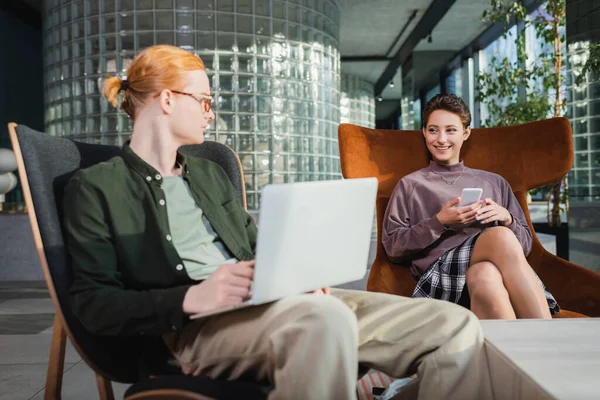 Smiling woman with cellphone looking at blurred boyfriend using laptop in hotel lobby — Stock Photo