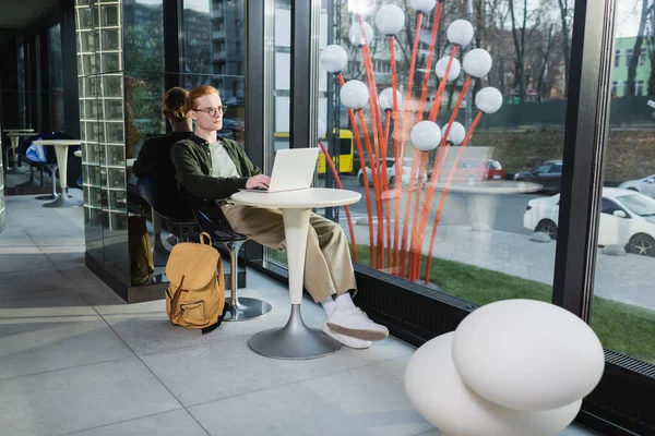 Redhead freelancer using laptop near backpack in hotel lobby — Stock Photo