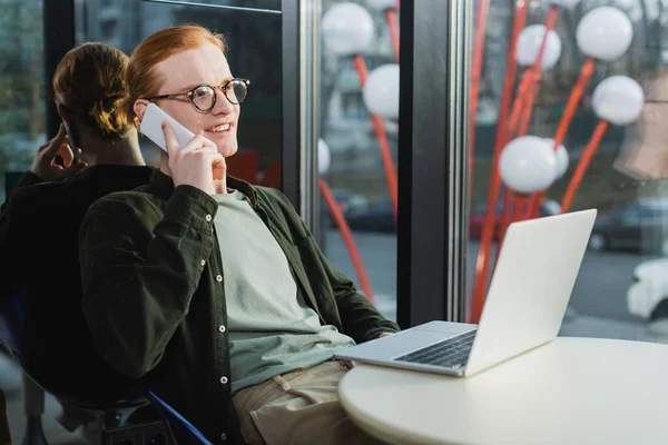 Happy red haired freelancer talking on smartphone near laptop in hotel lobby — Stock Photo