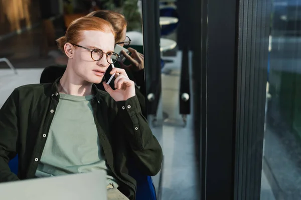 Homem ruivo falando no smartphone perto de laptop desfocado no lobby do hotel — Fotografia de Stock