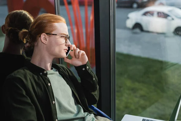 Vista lateral del joven freelancer hablando en smartphone cerca del portátil en el hotel - foto de stock
