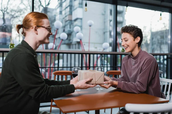 Smiling man giving present to girlfriend near wine in hotel cafe — Fotografia de Stock