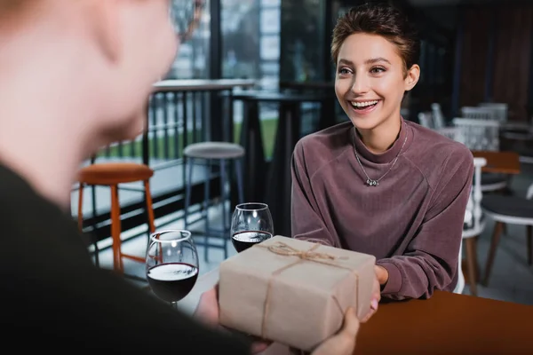 Excited woman holding present near wine and blurred boyfriend in hotel cafe — Fotografia de Stock