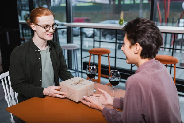 Smiling redhead man holding gift near blurred girlfriend and wine in hotel cafe — Stockfoto