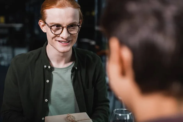 Smiling man in eyeglasses holding present near wine and blurred girlfriend in hotel — Foto stock