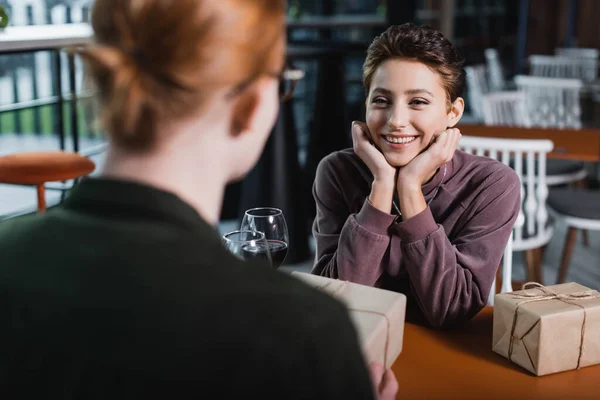 Joyful woman looking at blurred boyfriend with present near wine in hotel cafe — стоковое фото