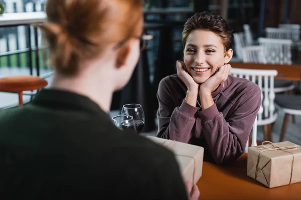 Joyful woman looking at blurred boyfriend with present near wine in hotel cafe — Stock Photo