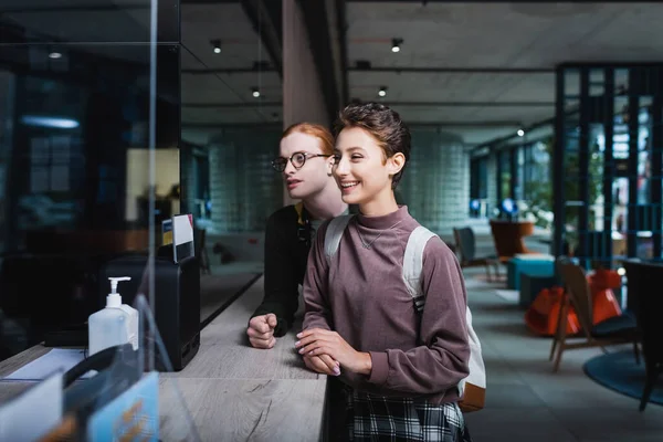 Positive woman with backpack standing near boyfriend on hotel reception — Stockfoto