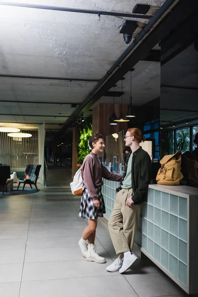 Side view of smiling couple with backpacks standing near reception of hotel — Fotografia de Stock