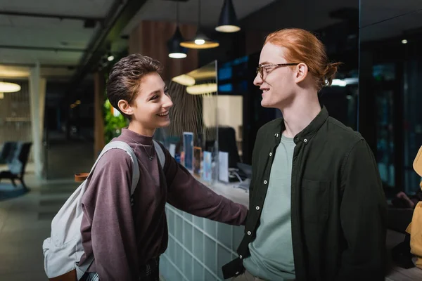 Smiling man talking to girlfriend with backpack near reception in hotel — Stockfoto