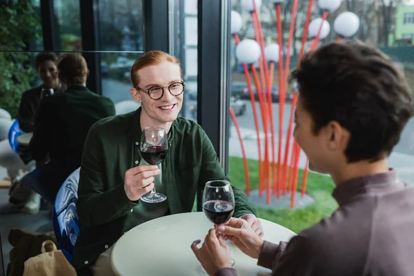 Smiling red haired man holding glass of wine near blurred girlfriend in hotel — Fotografia de Stock