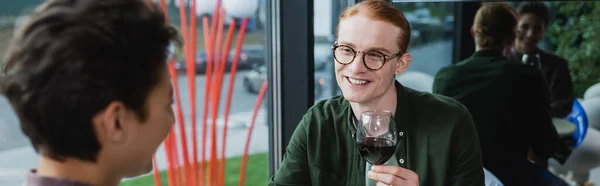 Smiling red haired man holding glass of wine near blurred woman in hotel, banner — стоковое фото