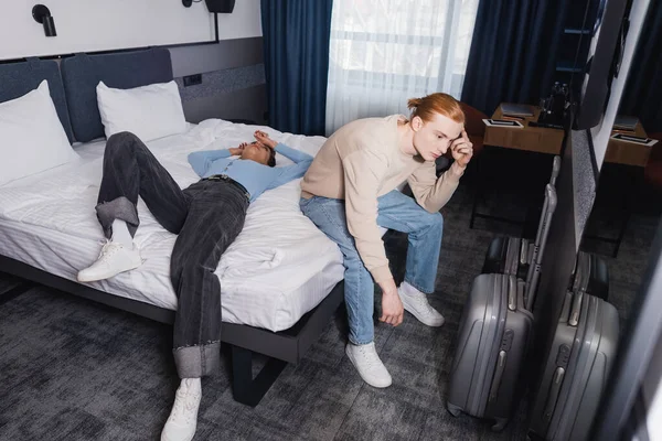 Cansado coupé sentado en la cama cerca de maletas en la habitación de hotel - foto de stock