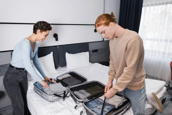 Vista lateral de la pareja sonriente desempacando maletas en la habitación del hotel - foto de stock