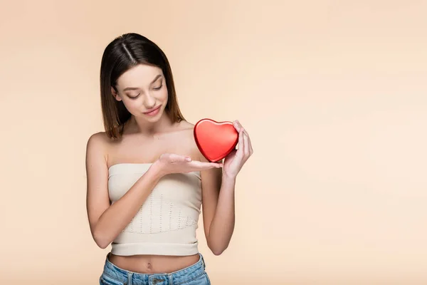 Mujer joven sosteniendo caja metálica roja en forma de corazón aislada en beige - foto de stock