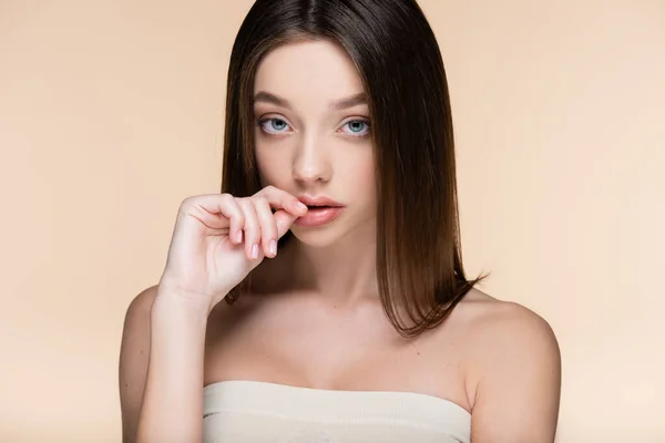 Pretty young woman with bare shoulders touching lips and looking at camera isolated on beige — Stock Photo
