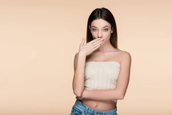 Giovane donna in crop top con spalle nude che coprono la bocca isolata sul beige — Foto stock