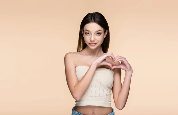 Smiling and pretty woman in crop top showing heart with hands isolated on beige — Foto stock