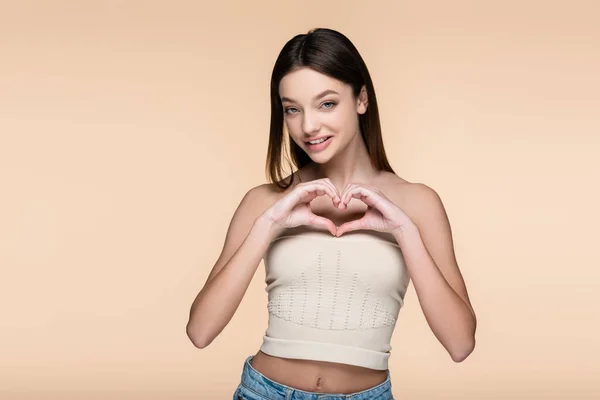 Cheerful young woman in crop top showing heart with hands isolated on beige — Stockfoto
