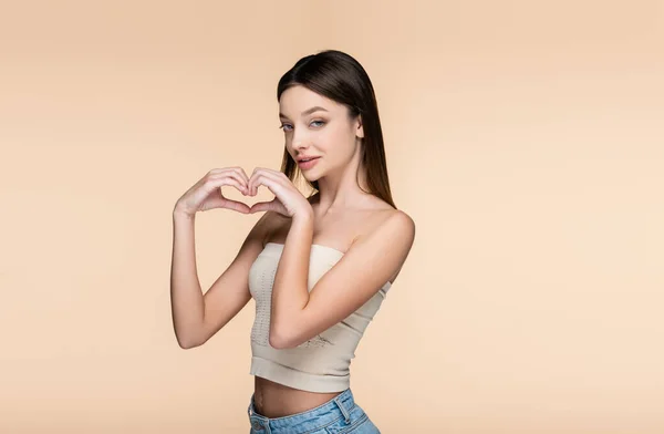 Brunette and pretty woman in crop top showing heart with hands isolated on beige — Stockfoto