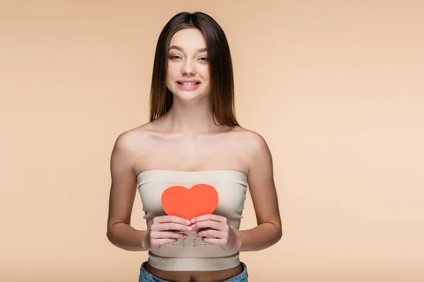 Happy woman in crop top with bare shoulders holding red paper heart isolated on beige — Stockfoto