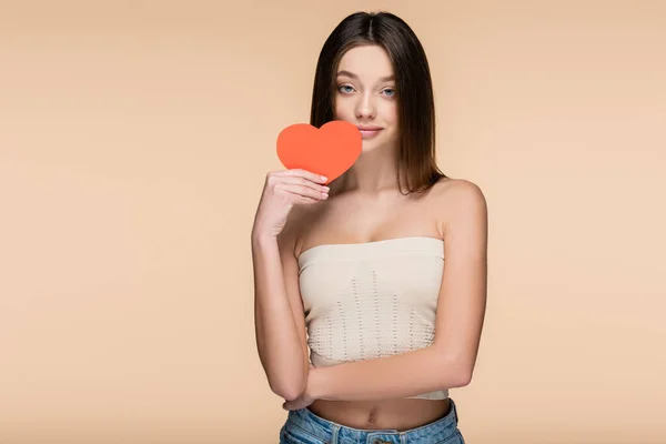 Mujer joven en la parte superior de la cosecha con hombros desnudos sosteniendo el corazón de papel rojo aislado en beige - foto de stock