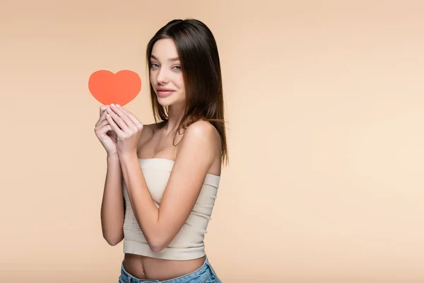 Mujer alegre en la parte superior de la cosecha con hombros desnudos sosteniendo el corazón de papel rojo aislado en beige — Stock Photo