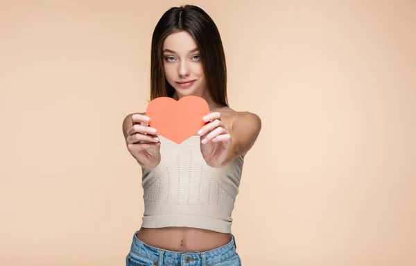 Brunette woman in crop top with bare shoulders holding red paper heart isolated on beige — Stock Photo