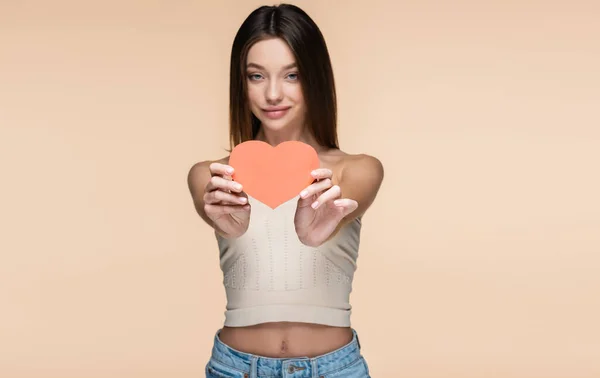 Smiling young woman in crop top with bare shoulders holding red paper heart isolated on beige — Stockfoto