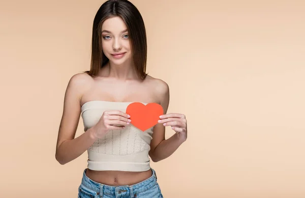 Young and cheerful woman in crop top with bare shoulders holding red paper heart isolated on beige — Stockfoto