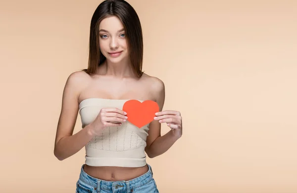 Mujer joven y alegre en la parte superior de la cosecha con hombros desnudos sosteniendo el corazón de papel rojo aislado en beige - foto de stock