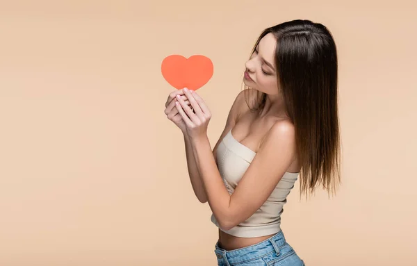 Mujer joven en la parte superior de la cosecha con los hombros desnudos mirando corazón de papel rojo aislado en beige - foto de stock